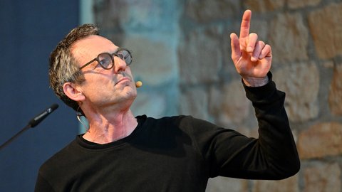 "Porträt eines Mannes mittleren Alters mit dunklem kurzen Haar und runder Brille. Er trägt ein schwarzes Langarmshirt und gestikuliert mit erhobener Hand nach oben. Der Hintergrund ist unscharfe blaue und beige Farbtöne. Ein Mikrofon ist sichtbar. Die Geste suggeriert eine ausdrucksstarke Sprechweise."