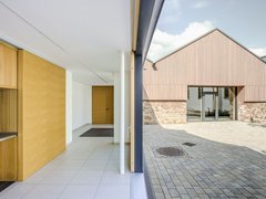 Das Foto zeigt eine Außenaufnahme eines modernen Gebäudekomplexes. Der Fokus liegt auf dem Übergang zwischen Innen- und Außenbereich.  Im Vordergrund steht eine geöffnete Schiebetür mit einer weißen Säule. Der Innenraum ist hell und schlicht gestaltet, mit weißen Wänden, hellgrauen Bodenfliesen und Holzakzenten. Links befindet sich ein Einbauschrank, während am Ende des Flurs eine geschlossene Holztür zu sehen ist.  Durch die geöffnete Tür blickt man auf einen gepflasterten Hof. Die Außenfassade des gegenüberliegenden Gebäudes besteht im unteren Bereich aus Natursteinen und im oberen Bereich aus einer senkrechten Holzverkleidung. Große Fenster und eine Glastür durchbrechen die Fassade. Ein Schachtdeckel ist im Pflaster sichtbar, und eine Bank steht im Hintergrund. Der Himmel ist leicht bewölkt.