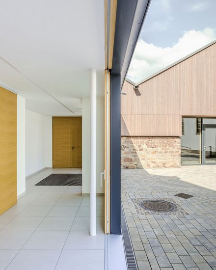 Das Foto zeigt eine Außenaufnahme eines modernen Gebäudekomplexes. Der Fokus liegt auf dem Übergang zwischen Innen- und Außenbereich.  Im Vordergrund steht eine geöffnete Schiebetür mit einer weißen Säule. Der Innenraum ist hell und schlicht gestaltet, mit weißen Wänden, hellgrauen Bodenfliesen und Holzakzenten. Links befindet sich ein Einbauschrank, während am Ende des Flurs eine geschlossene Holztür zu sehen ist.  Durch die geöffnete Tür blickt man auf einen gepflasterten Hof. Die Außenfassade des gegenüberliegenden Gebäudes besteht im unteren Bereich aus Natursteinen und im oberen Bereich aus einer senkrechten Holzverkleidung. Große Fenster und eine Glastür durchbrechen die Fassade. Ein Schachtdeckel ist im Pflaster sichtbar, und eine Bank steht im Hintergrund. Der Himmel ist leicht bewölkt.