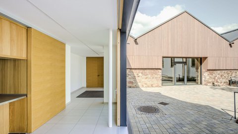 Das Foto zeigt eine Außenaufnahme eines modernen Gebäudekomplexes. Der Fokus liegt auf dem Übergang zwischen Innen- und Außenbereich.  Im Vordergrund steht eine geöffnete Schiebetür mit einer weißen Säule. Der Innenraum ist hell und schlicht gestaltet, mit weißen Wänden, hellgrauen Bodenfliesen und Holzakzenten. Links befindet sich ein Einbauschrank, während am Ende des Flurs eine geschlossene Holztür zu sehen ist.  Durch die geöffnete Tür blickt man auf einen gepflasterten Hof. Die Außenfassade des gegenüberliegenden Gebäudes besteht im unteren Bereich aus Natursteinen und im oberen Bereich aus einer senkrechten Holzverkleidung. Große Fenster und eine Glastür durchbrechen die Fassade. Ein Schachtdeckel ist im Pflaster sichtbar, und eine Bank steht im Hintergrund. Der Himmel ist leicht bewölkt.