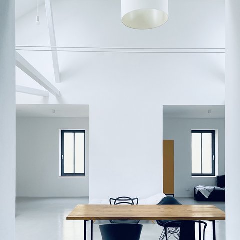 "Heller, minimalistischer Raum mit weißen Wänden und weißer Decke. Ein einfacher Holztisch aus hellem Holz mit schwarzem Stahlgestell steht in der Bildmitte. An der Decke eine große runde LED-Deckenleuchte. Im Hintergrund zwei offene Durchgänge zu weiteren Räumen mit dunkelblauen Türrahmen und Fensterrahmen. Links eine braune vertikale Blende oder Trennwand. Der Boden ist poliert grau."