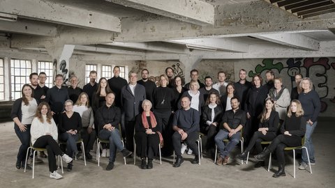 Großes Teamfoto mit ca. 30 Mitarbeitern in einer rustikalen Industriehalle mit Sichtbeton, Graffiti-Elementen und großen Fensterfronten
