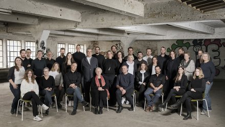 Großes Teamfoto mit ca. 30 Mitarbeitern in einer rustikalen Industriehalle mit Sichtbeton, Graffiti-Elementen und großen Fensterfronten