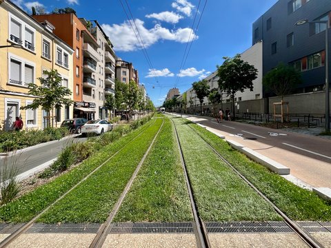 "Moderne urbane Straße mit begrünten Straßenbahngleisen in der Mitte. Links und rechts Wohngebäude in verschiedenen Farben: Gelb, Orange, Weiß und Grau. Grüne Bäume säumen die Straße. Ein weißes Auto parkt links. Mehrere Fußgänger sind auf der rechten Seite zu sehen. Der Himmel ist blau mit weißen Wolken. Stromkabel verlaufen über der Straße."