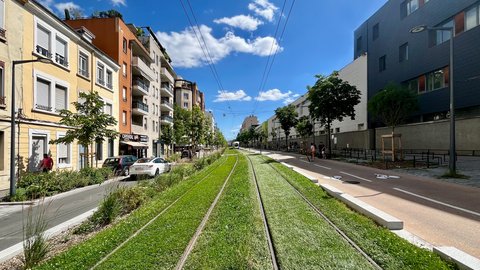 "Moderne urbane Straße mit begrünten Straßenbahngleisen in der Mitte. Links und rechts Wohngebäude in verschiedenen Farben: Gelb, Orange, Weiß und Grau. Grüne Bäume säumen die Straße. Ein weißes Auto parkt links. Mehrere Fußgänger sind auf der rechten Seite zu sehen. Der Himmel ist blau mit weißen Wolken. Stromkabel verlaufen über der Straße."