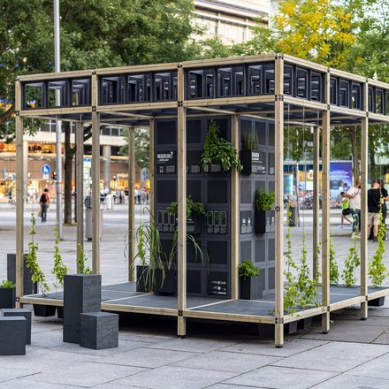 Auf einem städtischen Platz steht ein offenes Pavillon-Gerüst aus hellem Holz; in der Mitte ein schwarzer Modulturm mit Infotafeln und vielen kleinen Pflanzkästen, aus denen grünes Laub wächst. Daneben stehen niedrige schwarze Sitzquader; im Hintergrund Bäume und Passant:innen.
