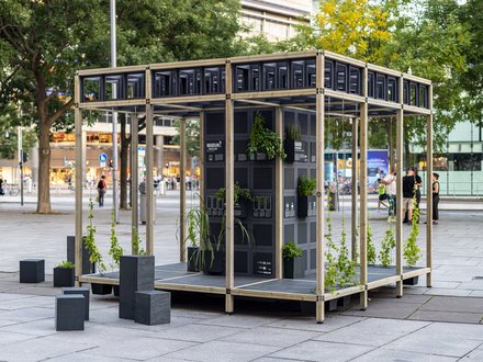 Auf einem städtischen Platz steht ein offenes Pavillon-Gerüst aus hellem Holz; in der Mitte ein schwarzer Modulturm mit Infotafeln und vielen kleinen Pflanzkästen, aus denen grünes Laub wächst. Daneben stehen niedrige schwarze Sitzquader; im Hintergrund Bäume und Passant:innen.