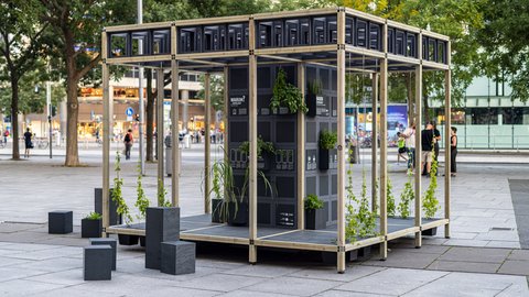 Auf einem städtischen Platz steht ein offenes Pavillon-Gerüst aus hellem Holz; in der Mitte ein schwarzer Modulturm mit Infotafeln und vielen kleinen Pflanzkästen, aus denen grünes Laub wächst. Daneben stehen niedrige schwarze Sitzquader; im Hintergrund Bäume und Passant:innen.