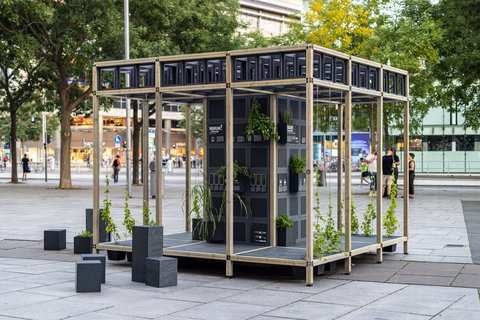 Auf einem städtischen Platz steht ein offenes Pavillon-Gerüst aus hellem Holz; in der Mitte ein schwarzer Modulturm mit Infotafeln und vielen kleinen Pflanzkästen, aus denen grünes Laub wächst. Daneben stehen niedrige schwarze Sitzquader; im Hintergrund Bäume und Passant:innen.