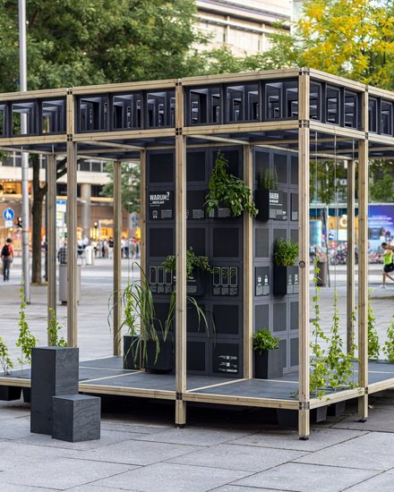 Auf einem städtischen Platz steht ein offenes Pavillon-Gerüst aus hellem Holz; in der Mitte ein schwarzer Modulturm mit Infotafeln und vielen kleinen Pflanzkästen, aus denen grünes Laub wächst. Daneben stehen niedrige schwarze Sitzquader; im Hintergrund Bäume und Passant:innen.