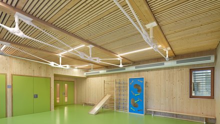Bewegungsraum: Grüner Boden, Holzdeckenlamellen und Turn‑/Spielgeräte; blaue Matte liegt aus.