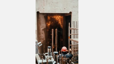 Baustelle in einem Betonschacht: Ein Arbeiter mit rotem Helm blickt auf eine dunkle Öffnung, in der bei Schneid- oder Schweißarbeiten helle Funken sprühen; ringsum Bewehrungsstäbe und Gerüstteile.