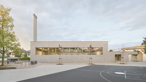 Flacher, moderner Schulgebäudekomplex mit großen Fenstern und zwei hohen Schornsteinen neben einem Schulhof mit Basketballfeld und Fahrradständern