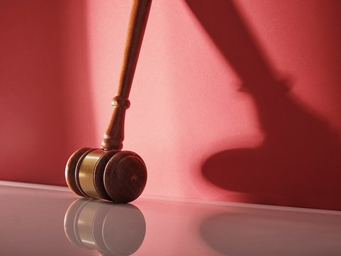 "Richterhammer mit Holzstiel und goldfarbener Metallbänderung liegt auf einer glänzenden, hellen Oberfläche vor einem rosa-roten Hintergrund. Der runde Hammerkopf wirft einen scharfen Schatten auf die Fläche. Das Bild symbolisiert Rechtsprechung und Justiz."