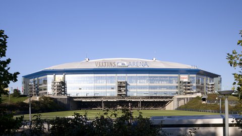 Außenansicht der Veltins-Arena in Gelsenkirchen an einem klaren, sonnigen Tag unter blauem Himmel. Die moderne Fassade des Stadions besteht hauptsächlich aus Glas und Stahl, darüber wölbt sich ein helles Dach. Der Schriftzug "VELTINS ARENA" ist groß oben in der Mitte der Fassade angebracht. Im Vordergrund sind Bäume und Sträucher zu sehen, dahinter ein grüner Rasen.