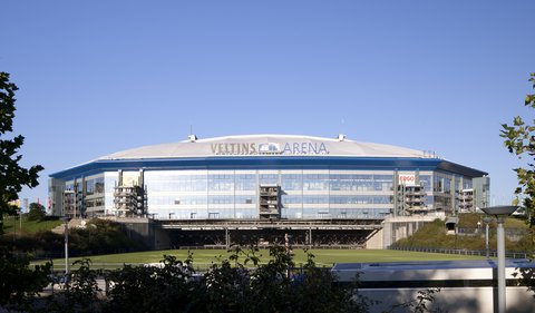 Außenansicht der Veltins-Arena in Gelsenkirchen an einem klaren, sonnigen Tag unter blauem Himmel. Die moderne Fassade des Stadions besteht hauptsächlich aus Glas und Stahl, darüber wölbt sich ein helles Dach. Der Schriftzug "VELTINS ARENA" ist groß oben in der Mitte der Fassade angebracht. Im Vordergrund sind Bäume und Sträucher zu sehen, dahinter ein grüner Rasen.