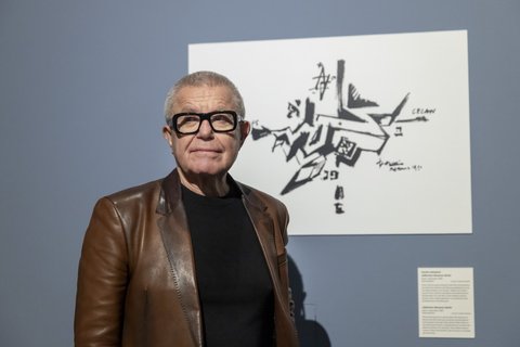 Älterer Mann mit Brille und brauner Lederjacke steht vor einer abstrakten schwarz-weißen Zeichnung an einer blauen Wand
