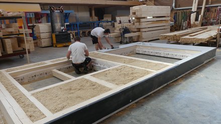 Zimmerei: Zwei Handwerker montieren einen Holzrahmenboden; Gefache sind mit Holzspänen als Dämmung gefüllt.