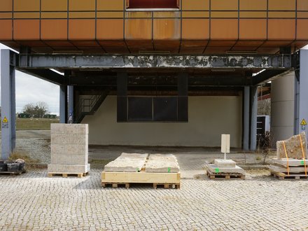 Unterseite eines erhöhten, orange verkleideten Gebäudes auf grauen Stahlstützen: Auf Kopfsteinpflaster stehen mehrere Paletten mit groben Stein- und Betonplatten, teils verzurrt; im Hintergrund eine Treppe und verwitterte Stahlträger bei bedecktem Himmel.