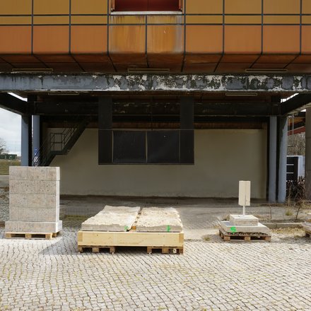 Unterseite eines erhöhten, orange verkleideten Gebäudes auf grauen Stahlstützen: Auf Kopfsteinpflaster stehen mehrere Paletten mit groben Stein- und Betonplatten, teils verzurrt; im Hintergrund eine Treppe und verwitterte Stahlträger bei bedecktem Himmel.
