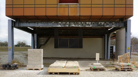 Unterseite eines erhöhten, orange verkleideten Gebäudes auf grauen Stahlstützen: Auf Kopfsteinpflaster stehen mehrere Paletten mit groben Stein- und Betonplatten, teils verzurrt; im Hintergrund eine Treppe und verwitterte Stahlträger bei bedecktem Himmel.