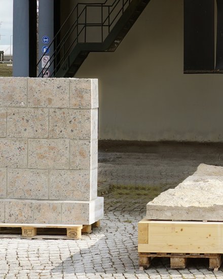 Unterseite eines erhöhten, orange verkleideten Gebäudes auf grauen Stahlstützen: Auf Kopfsteinpflaster stehen mehrere Paletten mit groben Stein- und Betonplatten, teils verzurrt; im Hintergrund eine Treppe und verwitterte Stahlträger bei bedecktem Himmel.