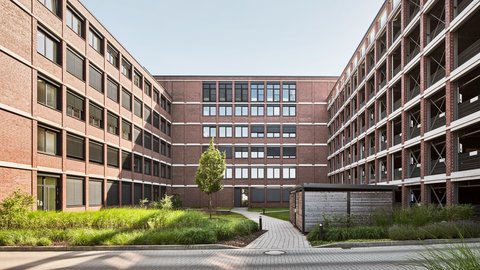 Innenhof mit gepflastertem Weg, mittig stehendem Baum und zwei mehrstöckigen Backsteingebäuden links und rechts