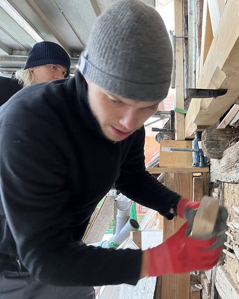 Zwei Personen in Mützen und rotem Handschuh arbeiten an einer alten Stein- und Holzwand