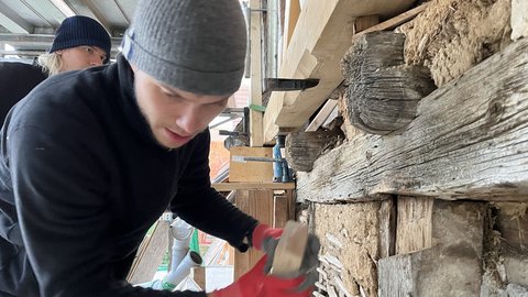 Zwei Personen in Mützen und rotem Handschuh arbeiten an einer alten Stein- und Holzwand