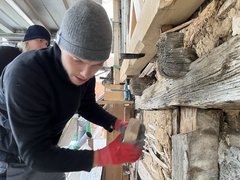 Zwei Personen in Mützen und rotem Handschuh arbeiten an einer alten Stein- und Holzwand