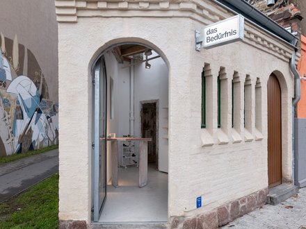 Kleines saniertes Eckgebäude mit Rundbogen-Eingang und Schild „das Bedürfnis“; Blick in einen minimalistischen Innenraum mit Ausstellungsmöbeln.