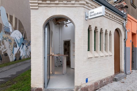 Kleines saniertes Eckgebäude mit Rundbogen-Eingang und Schild „das Bedürfnis“; Blick in einen minimalistischen Innenraum mit Ausstellungsmöbeln.