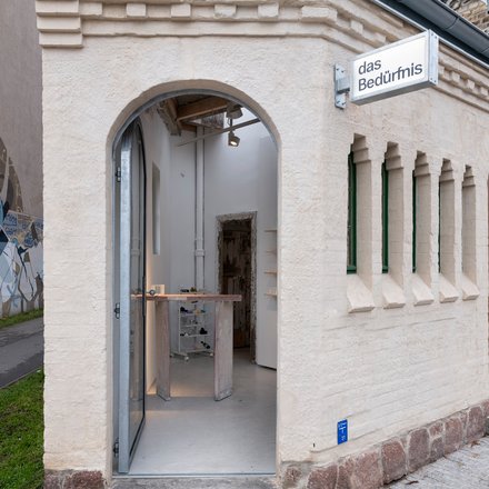 Kleines saniertes Eckgebäude mit Rundbogen-Eingang und Schild „das Bedürfnis“; Blick in einen minimalistischen Innenraum mit Ausstellungsmöbeln.
