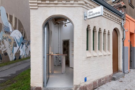 Kleines saniertes Eckgebäude mit Rundbogen-Eingang und Schild „das Bedürfnis“; Blick in einen minimalistischen Innenraum mit Ausstellungsmöbeln.