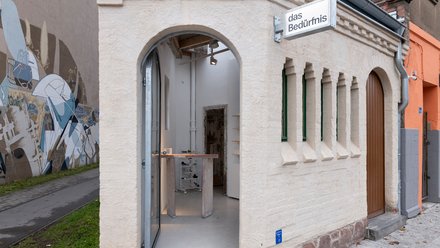 Kleines saniertes Eckgebäude mit Rundbogen-Eingang und Schild „das Bedürfnis“; Blick in einen minimalistischen Innenraum mit Ausstellungsmöbeln.