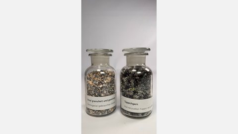 Zwei Glasgefäße mit Deckel, links gefüllt mit grauen und beigen Vinylgranulatstücken, rechts mit schwarzem Teppichgarn aus recycelten Fasern