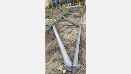 Das Bild zeigt eine Reihe von metallischen Stahlträgern, die auf einem unbefestigten, sandigen Untergrund ausgelegt sind. Die Träger sind geometrisch angeordnet und durch Querstreben miteinander verbunden, sodass eine tragende Struktur erkennbar wird. Im Hintergrund sind Baumaschinen und ein Gebäude zu sehen, was auf eine Baustellensituation hinweist. Die Stahlbauteile wirken gebraucht, aber funktionstüchtig und sind offensichtlich für eine erneute Montage vorbereitet. Zweck des Bildes ist es, die Wiederverwendung tragender Stahlbauteile im Bauprozess zu veranschaulichen und den praktischen Umgang mit ReUse-Materialien auf der Baustelle zu zeigen.