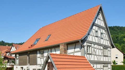 "Großes historisches Fachwerkhaus unter blauem Himmel mit roten Ziegeln auf steilem Dach. Fachwerk-Konstruktion mit dunklen Balken auf weißer Fassade. Mehrere Fenster und eine kleine moderne Veranda mit roten Dachziegeln im Vordergrund. Umgeben von Grünflächen mit Heckeneinfassung. Wooded hillside im Hintergrund."