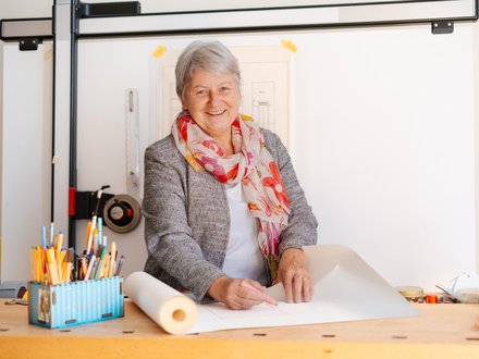 Lächelnde Frau mit grauen Haaren steht an einem Zeichentisch und arbeitet mit Bleistift an einer Bauzeichnung; auf dem Tisch liegen Zeichenwerkzeuge und Papierrollen.