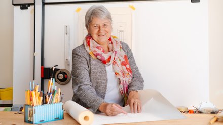 Lächelnde Frau mit grauen Haaren steht an einem Zeichentisch und arbeitet mit Bleistift an einer Bauzeichnung; auf dem Tisch liegen Zeichenwerkzeuge und Papierrollen.