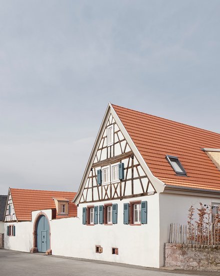 Weit geöffnete, hellblaue Torflügel in einem großen Sandsteinbogen; eine Frau und ein Mann stehen lächelnd in der geöffneten Türe und blicken in die Kamera. Kleineres Bild: Gesamtansicht eines Fachwerkhauses mit hohem Giebel und rotem Ziegeldach, davor niedrige weiße Mauer und ruhige Ortsstraße.