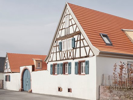 Weit geöffnete, hellblaue Torflügel in einem großen Sandsteinbogen; eine Frau und ein Mann stehen lächelnd in der geöffneten Türe und blicken in die Kamera. Kleineres Bild: Gesamtansicht eines Fachwerkhauses mit hohem Giebel und rotem Ziegeldach, davor niedrige weiße Mauer und ruhige Ortsstraße.