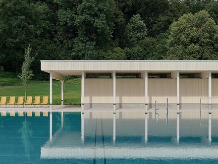 Modernes Freibad mit türkisblauem Becken im Vordergrund. Flaches, weiß-beiges Gebäude mit vertikalen Lamellen und weißen Stützen am Beckenrand. Links gelbe Liegestühle auf grünem Rasen. Im Hintergrund dichter grüner Laubwald unter grauem, bewölktem Himmel.