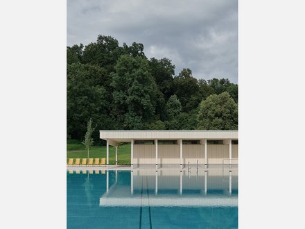 Modernes Freibad mit türkisblauem Becken im Vordergrund. Flaches, weiß-beiges Gebäude mit vertikalen Lamellen und weißen Stützen am Beckenrand. Links gelbe Liegestühle auf grünem Rasen. Im Hintergrund dichter grüner Laubwald unter grauem, bewölktem Himmel.