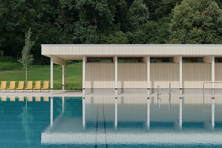 Modernes Freibad mit türkisblauem Becken im Vordergrund. Flaches, weiß-beiges Gebäude mit vertikalen Lamellen und weißen Stützen am Beckenrand. Links gelbe Liegestühle auf grünem Rasen. Im Hintergrund dichter grüner Laubwald unter grauem, bewölktem Himmel.