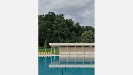 Modernes Freibad mit türkisblauem Becken im Vordergrund. Flaches, weiß-beiges Gebäude mit vertikalen Lamellen und weißen Stützen am Beckenrand. Links gelbe Liegestühle auf grünem Rasen. Im Hintergrund dichter grüner Laubwald unter grauem, bewölktem Himmel.