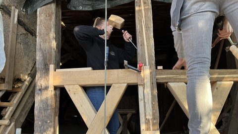 Zwei Personen arbeiten mit Hammer und Meißel an einer Holzkonstruktion in einem Dachstuhl