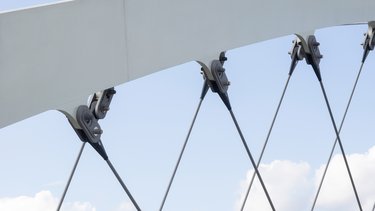 Detailansicht der Stahlkonstruktion einer Eisenbahnbrücke mit diagonal gespannten Zugstäben vor Himmel und Bäumen.
