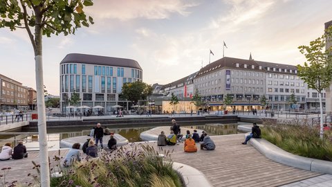eu gestalteter Bereich am Holstenfleet in Kiel mit Wasserlauf, Sitzstufen, bepflanzten Flächen und Aufenthaltsbereichen vor den umliegenden Gebäuden.