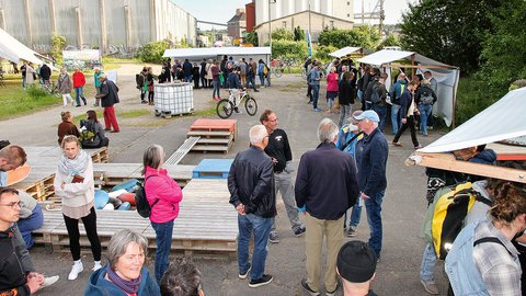 "Großes Sommerfest auf einem Industriegelände. Über hundert Menschen verschiedenen Alters verteilen sich auf einem Platz mit Betonboden. Im Vordergrund sitzen und stehen Besucher auf Holzpaletten-Sitzmöbeln mit farbigen Kissen. Im Hintergrund Zelte mit weißem Dach, ein Fahrrad, Getränkestand und Grünflächen. Links große Industriegebäude mit Wellblechdächern, rechts grüne Kletterpflanzen an der Fassade. Klarer Himmel. Die Atmosphäre wirkt gemütlich und gesellig."